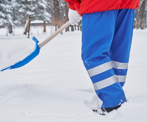 Snow removal on walkway in park. Concept of snow removal in winter with snow shovel. Janitor in blue pants and red jacket cleaning snow on walkway in park. Cleaner on outdoors close up, cropped.