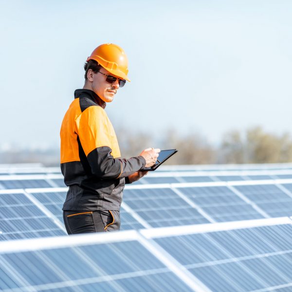Engineer in protective workwear carrying out service of solar panels with digital tablet on a photovoltaic rooftop plant. Concept of maintenance and setup of solar power station